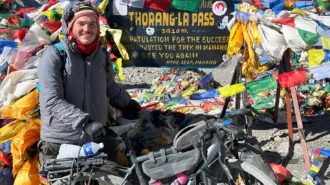 A man wearing a grey coat and black gloves is holding onto a bike and completing the Annapurna Circuit in Nepal.