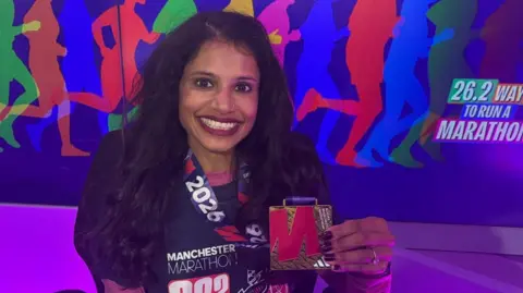 A South Asian woman wearing a purple Manchester Marathon 2026 medal as she smiles. Behind her is a hoarding with lots of runners in the background. 