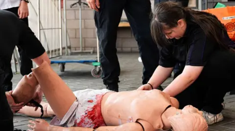 Northamptonshire Fire and Rescue Service Participants in a rescue training exercise use a casualty mannequin with simulated injuries while practising emergency response and patient handling techniques in an outdoor covered area.