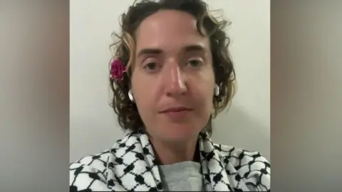 A woman with short hair sitting looking at the camera. She is wearing a keffiyeh, has earbuds in and has a flower above her ear. 