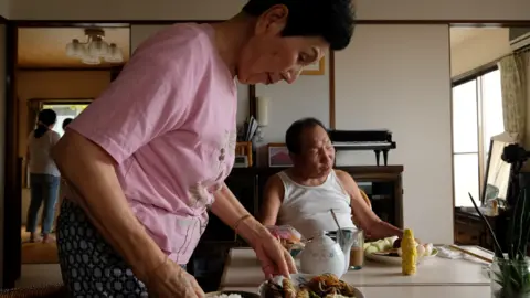 Seorang wanita berkemeja merah muda meletakkan makanannya di atas meja dapur sementara seorang pria dengan singlet putih duduk di ujung meja.