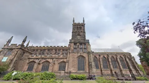A church building, set in grounds where there are trees and bushes. It has a tall tower with a clock and several ornate crenulations across its roof line.