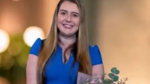 Hannah Fry, 29, who has long, straight blonde hair and is smiling at the camera while wearing a matching blue blouse and skirt and holding a bouquet of pink and yellow roses. 