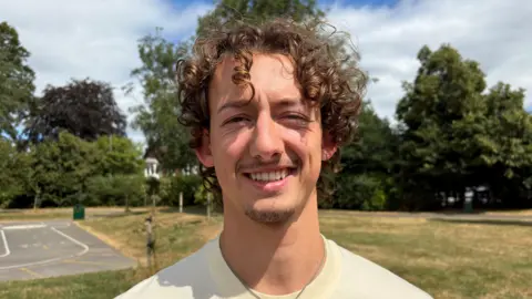 Jack has curly brown hair and short stubble. He smiles at the camera, standing in a park with grass and trees behind him. He wears a yellow t-shirt. 