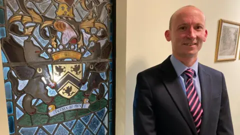 A man is dressed smartly, in a suit and tie. He is standing next to a stained glass window.
