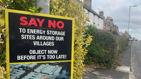 A large sign saying 'Say No to Energy Storage' at the entrance to a village street