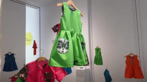 Exhibit of brightly-coloured children's dresses on clothes hangers hanging from the ceiling, set against white walls