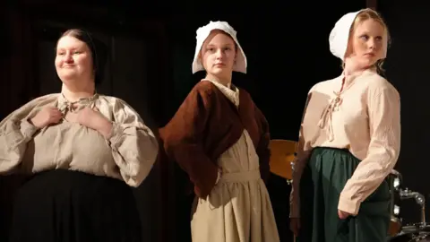 Red Rose Chain Three women in Tudor dress are standing on a stage, the middle one looks directly at the camera, the other two look out to each side.