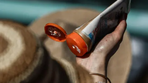EPA A hand clasping an open orange sun cream bottle , ready to apply it. The packaging is brown and white and the lid is orange. A straw sun hat can also be seen in the photo. 
