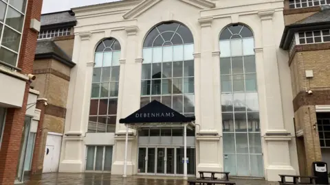 The large, vacant Debenhams building. It is three levels tall and has large, archway-shaped windows split between cream pillars. The building faces on to an outdoor shopping street. It is pictured on a rainy day.