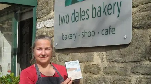 Hannah Parnell A woman in a red T shirt and apron holds a small booklet standing in front of a shop sign that reads 'two dales bakery'