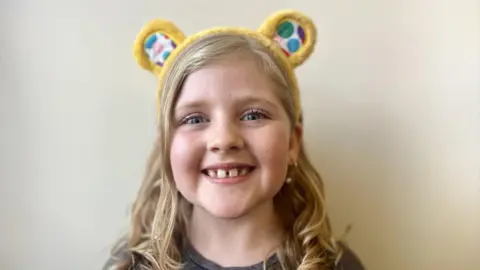 A young girl smiles at the camera. Her eyes are blue and her hair is blonde and curly. She is wearing BBC Children in Need Pudsey ears. The background is a cream colour. She has a grey t-shirt on, although only the top of her shoulders can be seen - the photo is a headshot.