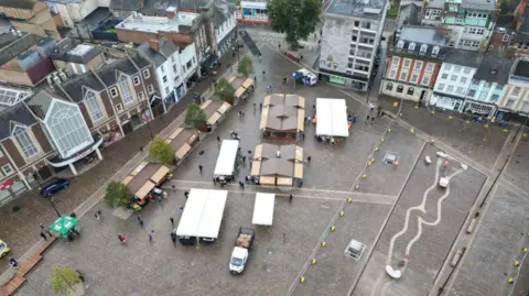 BBC An aerial view of Northampton Market Square. The square is surrounded by tall Victorian buildings. There are hard-standing covered stalls with roofs in the square. It is a dull day.