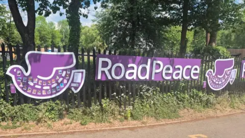Road Peace A large purple sign with Road Peace in white lettering hangs on a metal fence with trees behind. There are two purple doves, one on either side, with photos attached to them.