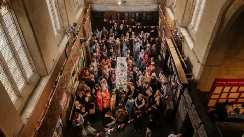 Shot from above depicting a 1940s railway station set created in the chancel gallery at the 20-21 Arts Centre. There are dozens of people in costume looking up towards the camera, along with bunting and people sat at a table eating food.

