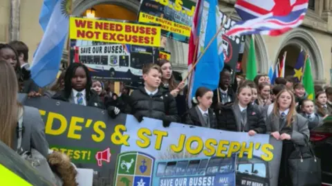 Pupils in school uniform hold up banners and flags. One banner reads "we need buses, not excuses" while another says "stop brutal school journey cuts".