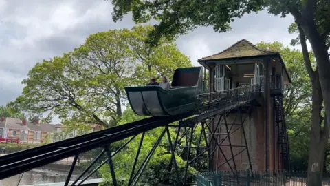 BBC The red brick tower of the splash boat ride has a tiled roof and a metal flume extending down into the lake. The green boat is halfway down the slide and two figures are enjoying the ride