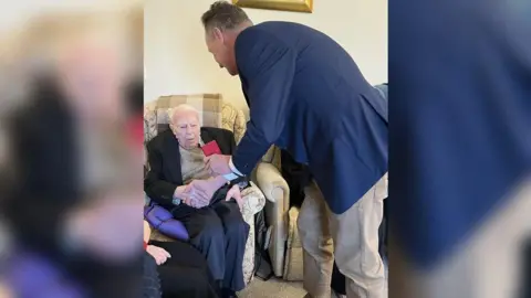 Sarah Dyke John sitting in an armchair being presented with a red wallet containing the medal by another man. Both are dressed smartly in suit jackets