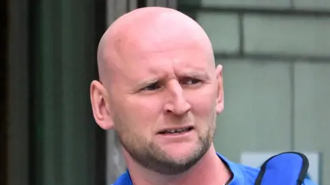 A bald headed man stands in front of a green wall. he has a bag over his shoulder and is wearing a blue top