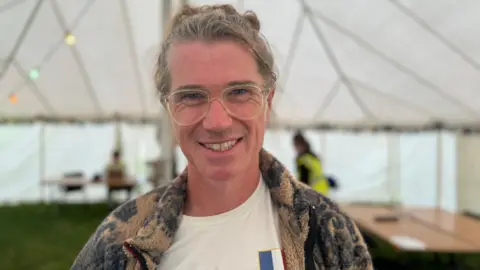 A man with grey/brown hair tied in a bun, smiles at the camera. He is wearing glasses and a brown jacket with grey patterns. He is standing inside a large white tent.