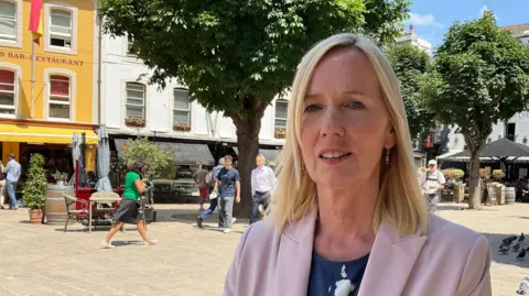 Catherine is wearing a light pink blazer and patterned navy top stands in a sunny town square. Behind her are colourful buildings, outdoor restaurant seating, several people walking, leafy trees, and a group of pigeons near a stone statue.