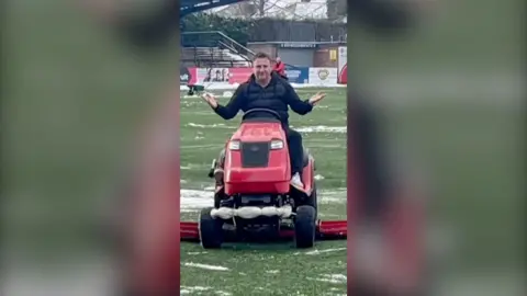 Rob Smethurst has dark hair and is wearing a black puffer jacket. He is sat on a red lawnmower on a football pitch.