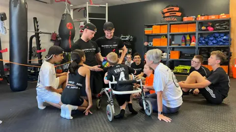 A group of children with Bobby and Shay looking at a multicoloured wheel. They are in the gym with punchbags in the background and apparatus. There is a woman wearing a white t-shit which is Bobby's nan.
