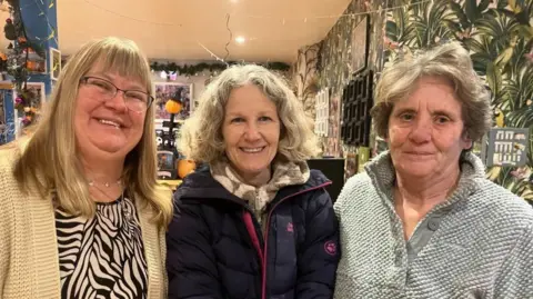 Vikki Irwin/BBC (left to right) Deidre Charleston, Loraine Cuzner and Gill Lewis are standing together closely and smiling at the camera. They are standing in front of a wall decorated with a leafy green wallpaper. Deidre on the left is wearing a light-coloured cardigan over a zebra-print top. Loraine in the middle is dressed in a dark puffer jacket. Gill on the right is wearing a textured light grey jumper.