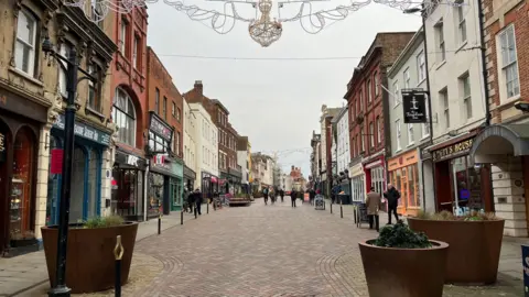 Gloucestershire County Council A cobbled street with shops on either sides and bollards separating the pavement from the road.