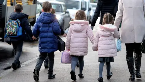 PA Media Five superior   schoolhouse  property  children walking connected  a street. There are 3  boys, wearing bluish  jackets, and 2  girls wearing pinkish  jackets. The girls are holding hands, and 1  of the girls is holding hands with an big  who is besides  wearing a pinkish  jacket.