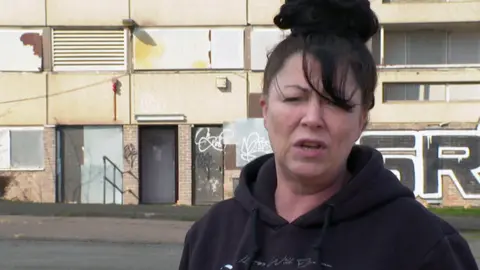 Head and shoulders view of a woman with dark hair and a dark hoodie standing in front of the flats, which are covered in graffiti