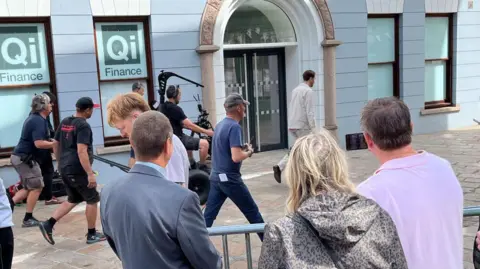 A film crew in the town of St Helier in Jersey watched by members of the public and police. An actor walks down a shopping street, followed by various members of the crew including a mounted camera operator.