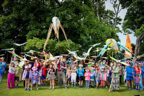 Fringe by the Sea People hold sticks with paper structures of colourful crafted birds. The group are standing in a crowd on the grass in front of some trees. The ages range from young children up to adults. 