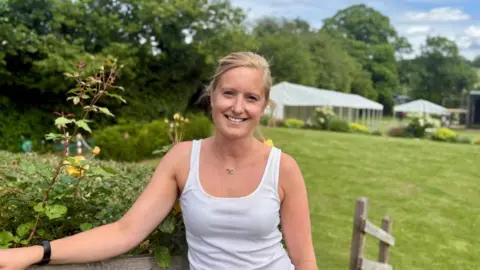 A woman with tied back blond hair, wearing a white vest top stands in a manicured garden with a large gazebo in the back ground.