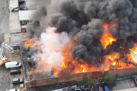 Bedfordshire Fire and Rescue Service A large single storey building has a raging orange fire inside and thick black smoke rising from it.