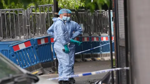 Two forensic investigators wearing light blue protective suits, face masks, and blue hairnets stand on a pavement behind blue and white police cordon tape. Blue road barriers are visible in the background.