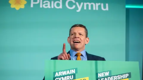 Matthew Horwood Rhun ap Iorwerth, a man with short brown hair in a navy suit, speaks while standing at a lectern.