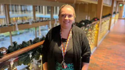 BBC / Gem O'Reilly Amy Brown, who is wearing a black top and blouse, is standing in front of a handrail on a walkway indoors. She is looking at the camera and smiling. The handrail is decorated with Christmas decorations.