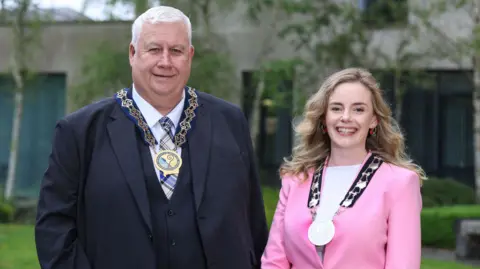 NMANDD Glyn Hanna and Doire Finn are standing next to each other and smiling. Both are wearing their chains of office. Hanna is wearing a navy suit and Finn is wearing a pink suit. 