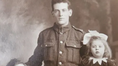 Neil Anderson A black and white photograph of a young man in uniform with his arm around a child with a bow in her hair and a person sitting in front of him.