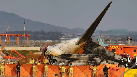The charred tail of Jeju Air Flight 2216 surrounded by firefighters at the end of the runway in Muan Airport in south-west South Korea