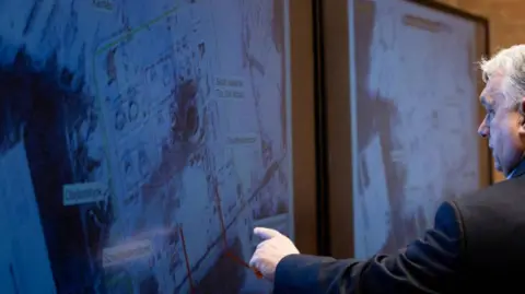 Hungarian government Viktor Orban pointing to a satellite image showing some red lines with his left-hand finger 