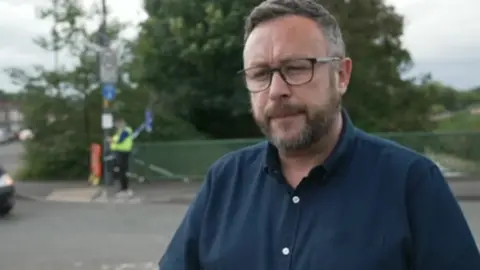 A man with a beard and glasses, wearing a blue shirt looks at the camera. Behind him is a canal bridge and a policeman standing next to a cordon