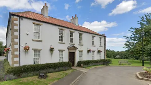 Shotton Hall. It is a two-storey white country house, surrounded by flowers and shrubs.