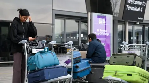 Two people outside Terminal 4 at London Heathrow Airport in London, on Friday 21 March London's with trolleys full of luggage. One person is looking at her phone while the other is sitting down. Heathrow airport suffered its worst disruption in at least two decades, after a nearby fire cut power to the hub and brought travel to a standstill for hundreds of thousands of passengers. 