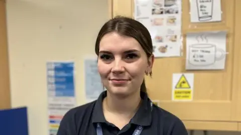 Shaun Whitmore/BBC A woman with dark hair that is tied back smiles at the camera. She has small gold hoop earrings in. She is wearing a black polo top with a lanyard around her neck.