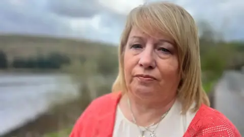 Maxine Johnson has blonde hair. She is looking directly into the camera, and is wearing a white t shirt and a pink cardigan