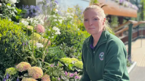Karen is crouching next a green flowerbed mixed with white and purple flowers. She is wearing a dark green zip up jumper with a Wimbledon emblem on the left side of the jumper. She has light ginger hair swept back into a ponytail. She's smiling at the camera.