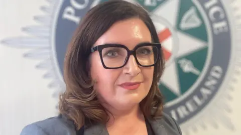 A woman with black glasses wearing a grey blazer. She has brown hair.