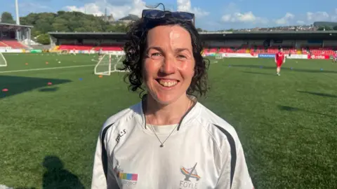 BBC Meabh O'Neill stands in the middle of a football pitch. An empty stand filled with red seats is in the background.  A lone figure dressed in a red kit walks towards her. The woman is standing in front of a set of goals and is wearing a white football shirt with the words Foyle pride written on it.